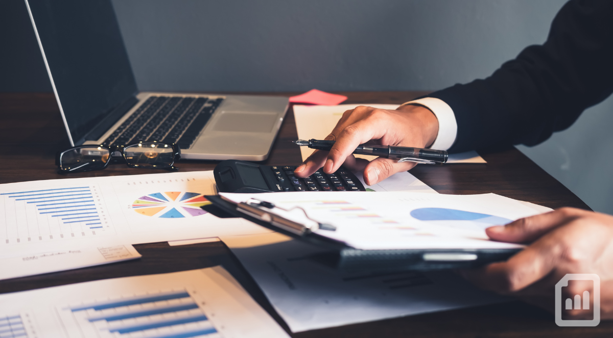 close-up of a desk with financial charts, laptop, and someone's hand holding a pen